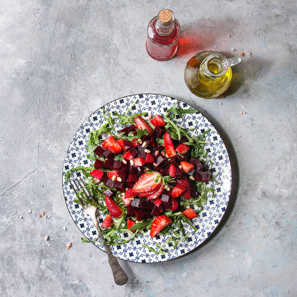 Beetroot and strawberry salad served with arugula and nuts on ceramic plate with bottles of fruit ocet and olive oil over grey texture background. Top view, copy space. Healthy eating - Valokuva, kuva
