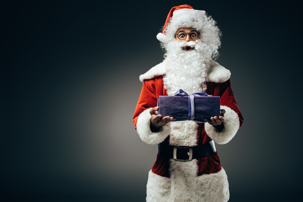 surprised santa claus in costume holding gift box isolated on grey background  - Foto, Imagen