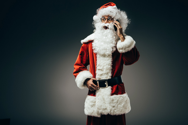 Schockierter Weihnachtsmann im Kostüm spricht auf Smartphone isoliert auf grauem Hintergrund  - Foto, Bild