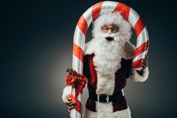sorprendido santa claus en traje de pie con gran palo de Navidad rayado aislado sobre fondo gris
  - Foto, Imagen