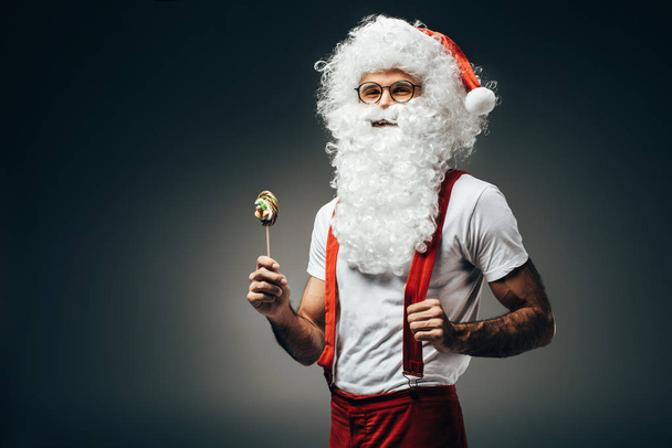 smiling santa claus in eyeglasses standing with colorful lollipop isolated on grey background  - Fotografie, Obrázek