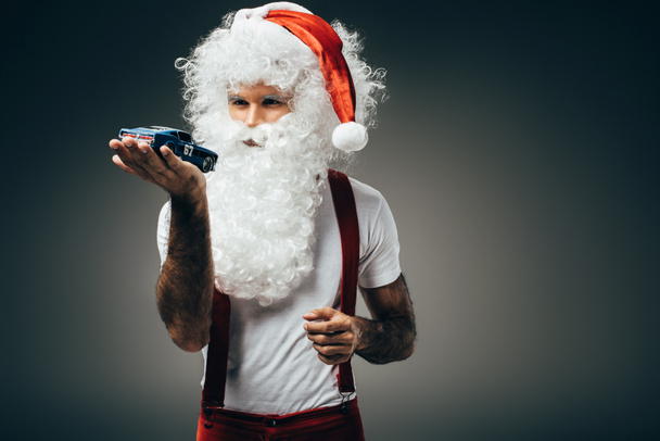 serious santa claus holding toy car on hand palm and standing isolated on grey background  - Zdjęcie, obraz