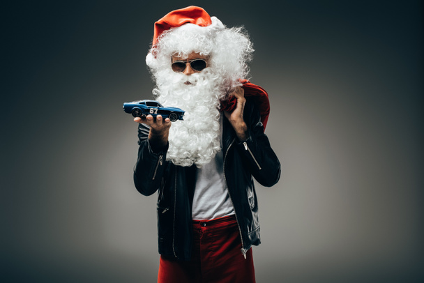 Άγιος Βασίλης σε γυαλιά ηλίου και δερμάτινο σακάκι κρατώντας παιχνίδι αυτοκίνητο και Χριστουγεννιάτικο σάκο απομονώνονται σε γκρι φόντο  - Φωτογραφία, εικόνα