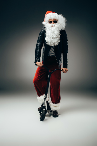 seriöser Weihnachtsmann in Sonnenbrille und Lederjacke mit Tretroller auf grauem Hintergrund  - Foto, Bild