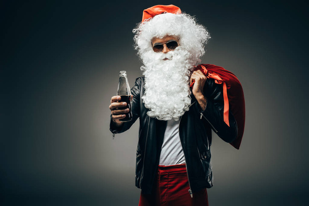 κομψό Αϊ Βασίλη με γυαλιά ηλίου και δερμάτινο σακάκι δείχνει μπουκάλι σόδα κρέμα και κρατώντας Χριστούγεννα τσάντα πάνω από τον ώμο που απομονώνονται σε γκρι φόντο  - Φωτογραφία, εικόνα