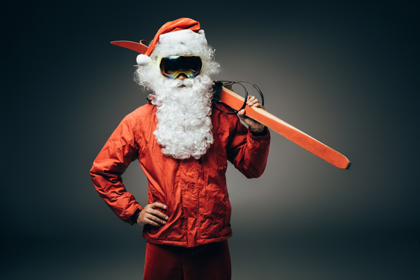 santa claus in ski mask and windbreaker holding skis over shoulder and standing with hand on waist isolated on grey background  - Photo, Image