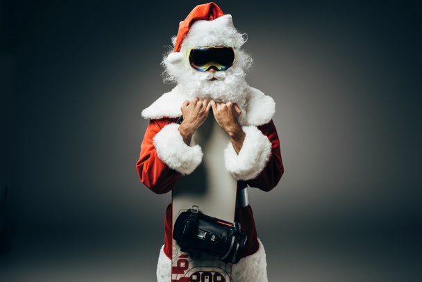 santa claus in ski mask standing with snowboard isolated on grey background  - Photo, Image