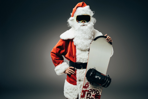 luottavainen joulupukki hiihtomaskissa seisoo käsi vyötäröllä ja pitää lumilauta eristetty harmaalla pohjalla
  - Valokuva, kuva