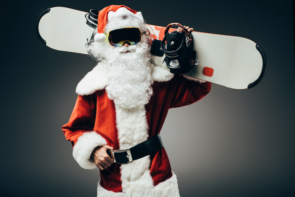 serious santa claus in ski mask standing with snowboard over shoulder isolated on grey background  - Photo, Image