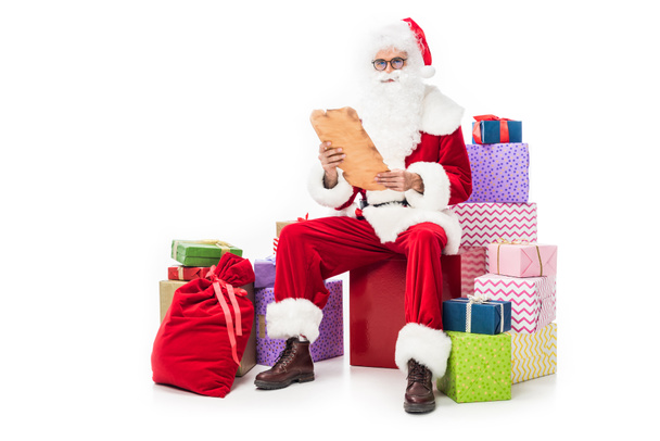 santa claus in eyeglasses reading old parchment and sitting on pile of gift boxes isolated on white background  - Zdjęcie, obraz