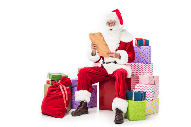 шокированный Санта Клаус в очках, читающий старый пергамент и сидящий на куче подарочных коробок, изолированных на белом фоне
  - Фото, изображение
