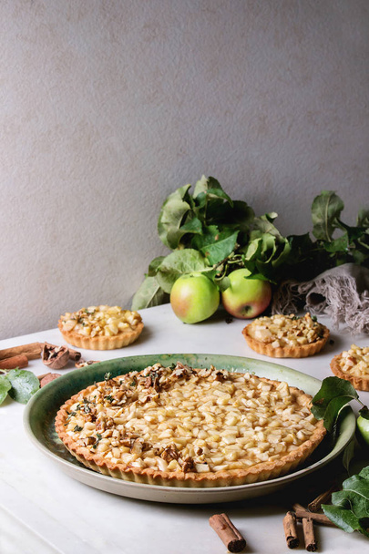 Homemade sweet apple shortbread tart in green ceramic plate and tartlets with cinnamon sticks, walnuts, apples branches above on white marble table. Autumn baking. - Foto, Imagem