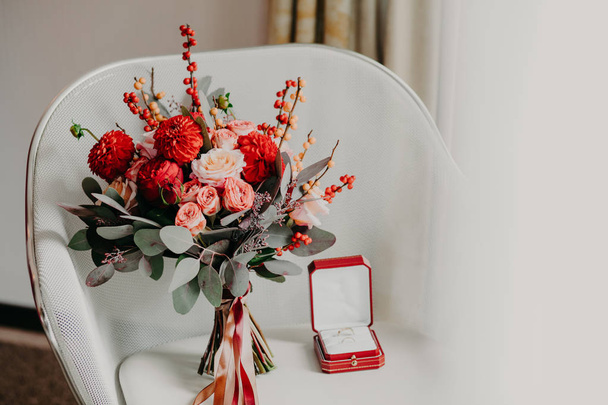 Foto horizontal de hermoso bouguet y anillos de boda de oro en caja roja en sillón blanco en habitación acogedora. Concepto de compromiso. Preparación de boda. Celebración
 - Foto, imagen