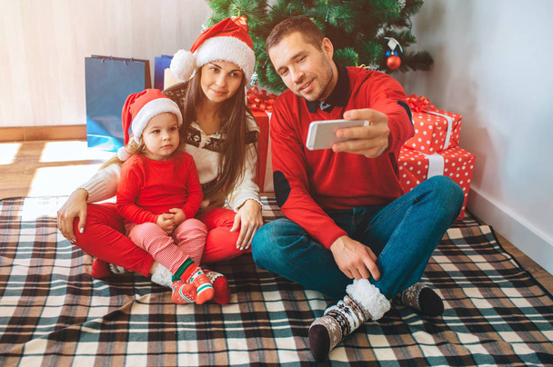 Merry Christmas and Happy New Year. Young man sits besides woman and child. He holds phone and takes selfie. Woman and kid lok at it and pose. Family wears Christmas clothes and hats. - Fotó, kép