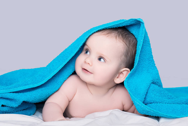 Surprised smiling baby girl or boy with big blue eyes lying on belly under the blue towel after shower, isolated over grey background - Fotografie, Obrázek