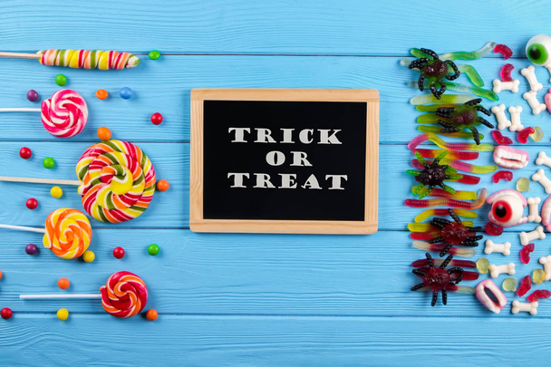 Trick or treat text written on blackboard with decorative paper bats, eyeball shaped candy, gummy worms, spiders & bones. Halloween decor concept. Background, copy space, close up, top view, flat lay. - Photo, image