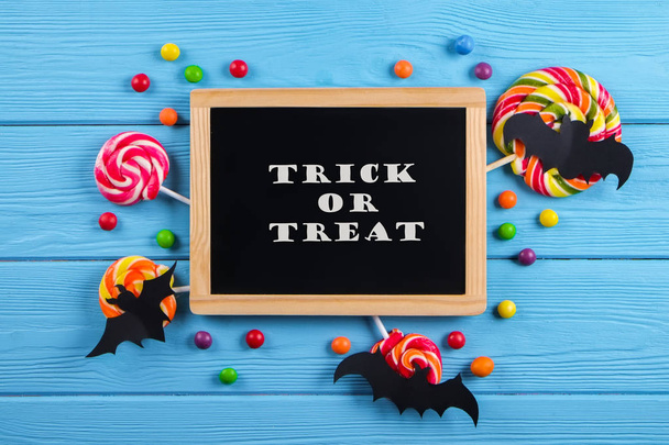 Trick or treat text written on blackboard with decorative paper bats, eyeball shaped candy, gummy worms, spiders & bones. Halloween decor concept. Background, copy space, close up, top view, flat lay. - Photo, Image