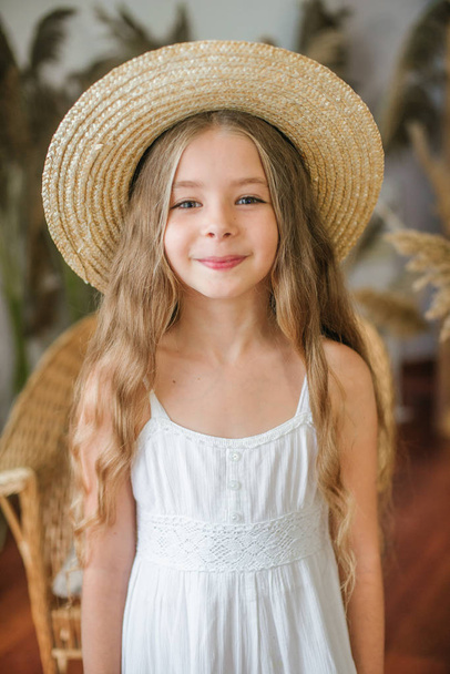 A sweet little girl with long blond hair in a white sarafan and a straw hat in a rattan chair and a reed decor - Foto, immagini