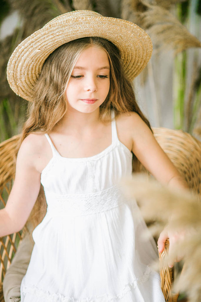 A sweet little girl with long blond hair in a white sarafan and a straw hat in a rattan chair and a reed decor - Foto, imagen