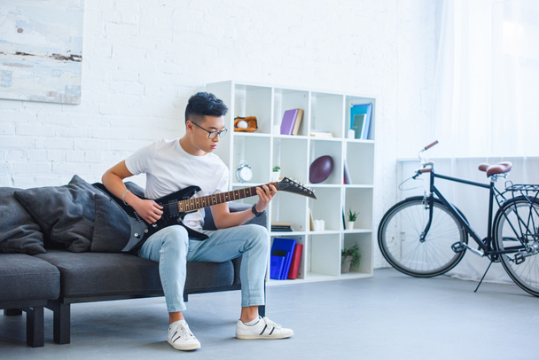 handsome asian man playing black electric guitar on sofa at home - Fotoğraf, Görsel