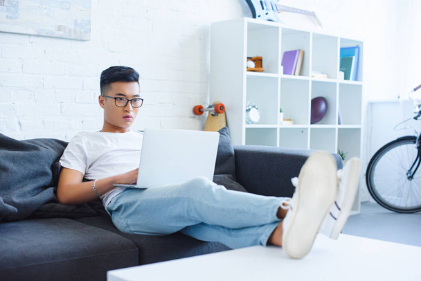 döbbenve csinos ázsiai ember használ a laptop otthon a kanapén - Fotó, kép