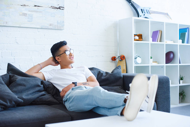 smiling handsome asian man sitting on sofa with legs on table at home, looking away - Photo, image