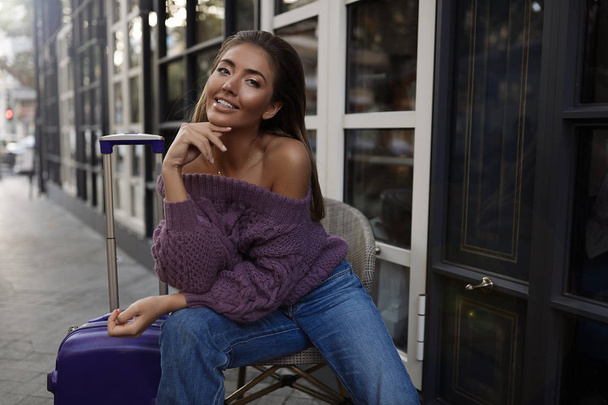 Beautiful female with the blue suitcase sitting on the chair near the cafe, in the purple pullover,jeans, makeup,hair style,  autumn, blonde, happy, knitted, smiling, hands near the face - Photo, Image