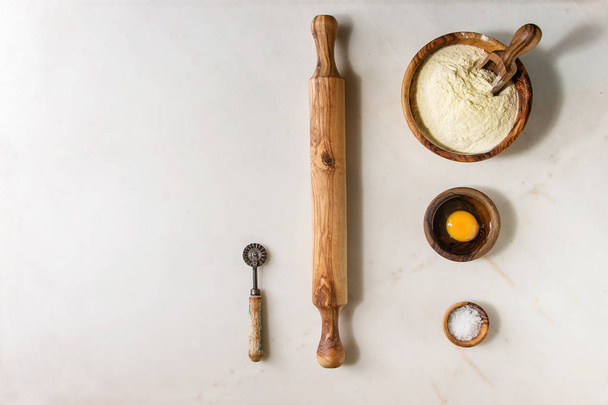 Ingredients for homemade italian pasta ravioli cooking semolina flour, egg yolk, sea salt with olive wood utensils bowls, scoop and rolling pin over white marble background. Flat lay, space - Foto, imagen