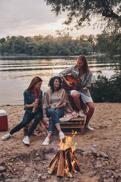 young people in casual wear enjoying camping near lake at sunset, man playing guitar  - Zdjęcie, obraz