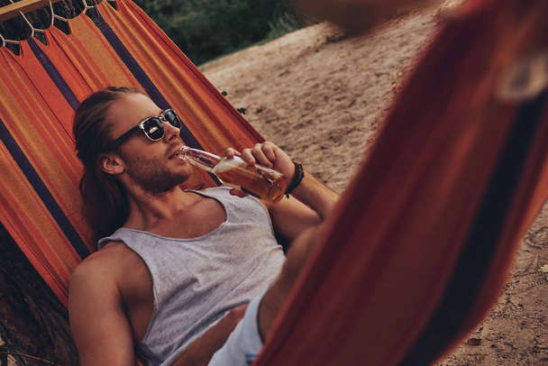 handsome stylish Caucasian man in sunglasses drinking beer while lying in hammock - Foto, imagen
