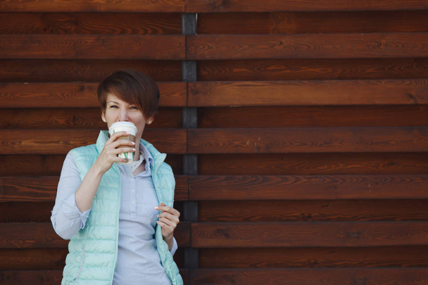 Młoda ładna kobieta w Błękitnej Kurtki jest picie kawy ze szklanki i patrzy na aparat, uśmiechając się na brązowym tle drewnianych. - Zdjęcie, obraz