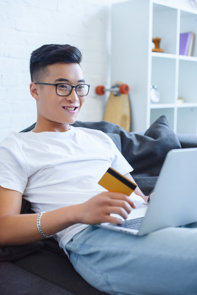 Улыбающийся красивый азиатский мужчина с ноутбуком и с кредиткой на диване дома
 - Фото, изображение