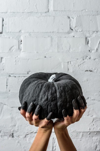 cropped shot of woman holding black painted halloween pumpkin in front of white brick wall with hands - Foto, Bild