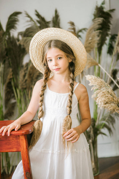 A sweet little girl with long blond hair in a white sarafan and a straw hat in a rattan chair and a reed decor - Fotografie, Obrázek