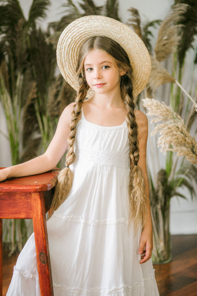 A sweet little girl with long blond hair in a white sarafan and a straw hat in a rattan chair and a reed decor - 写真・画像