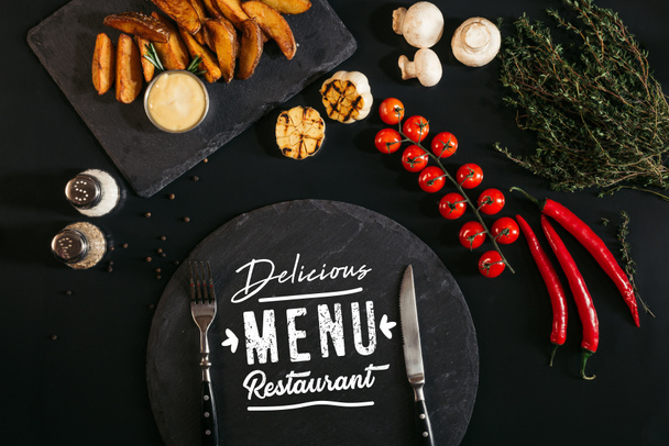 vista dall'alto di lavagna con scritta "delizioso ristorante menu" e forchetta e coltello, patate al forno, spezie e verdure su nero
    - Foto, immagini