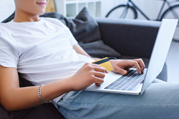 cropped image of man in casual clothes using laptop and holding credit card on sofa at home - 写真・画像