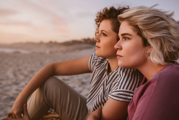 young lesbian couple sitting side by side together on a sandy beach watching a romantic sunset - 写真・画像