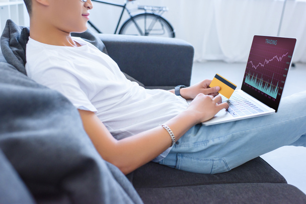 geschnittenes Bild eines Mannes, der Laptop mit Handelsgerät benutzt und Kreditkarte auf dem heimischen Sofa hält - Foto, Bild