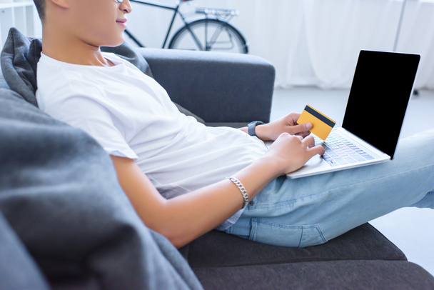 image recadrée de l'homme en utilisant un ordinateur portable avec écran vierge et tenant la carte de crédit sur le canapé à la maison
 - Photo, image