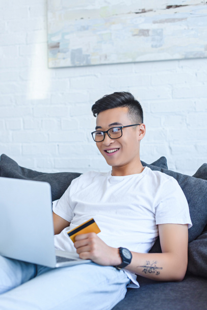 smiling handsome asian man using laptop on sofa and holding credit card at home - Foto, imagen