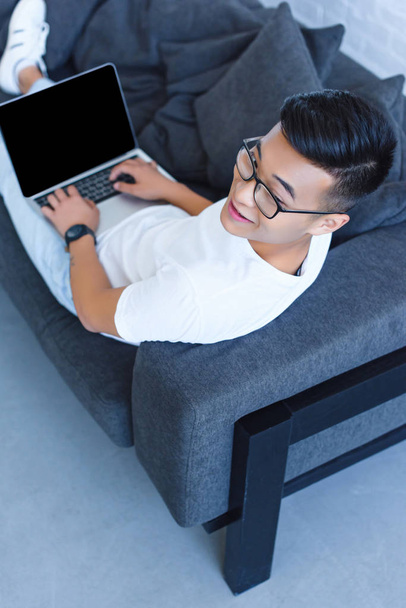 high angle view of handsome asian man lying on sofa and using laptop with blank screen at home - Photo, Image