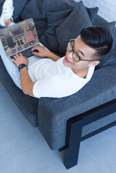 vista de ángulo alto de hombre asiático guapo acostado en el sofá y el uso de ordenador portátil con aparato de reserva en línea en casa
 - Foto, Imagen