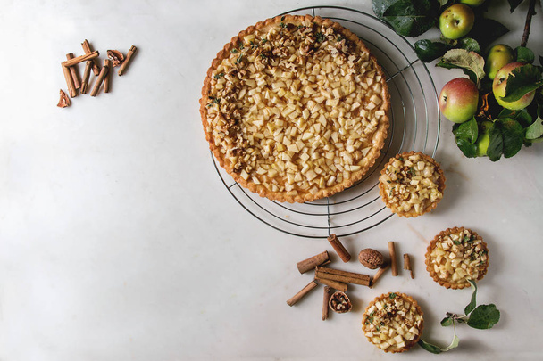 Homemade sweet apple shortbread tart on cooling rack and tartlets with cinnamon sticks, walnuts, apples branches above on white marble background. Autumn baking. Flat lay, space - Valokuva, kuva