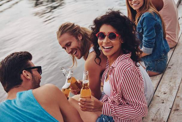 laughing young people in casual wear smiling and holding beer while sitting on pier at lake water  - Foto, Bild