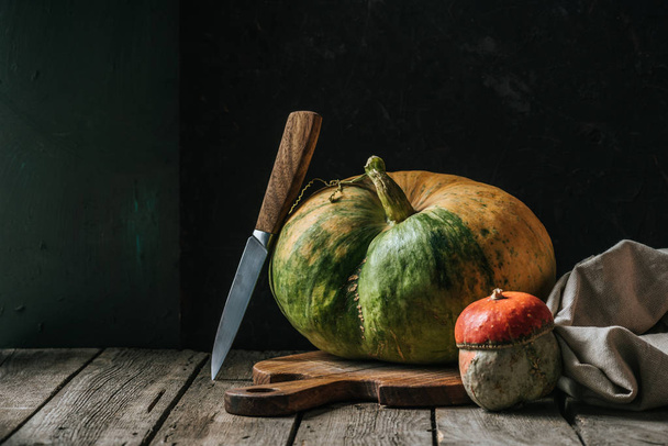 close up view of food composition with pumpkins, knife and cutting board arranged on wooden surface on dark background - Фото, изображение