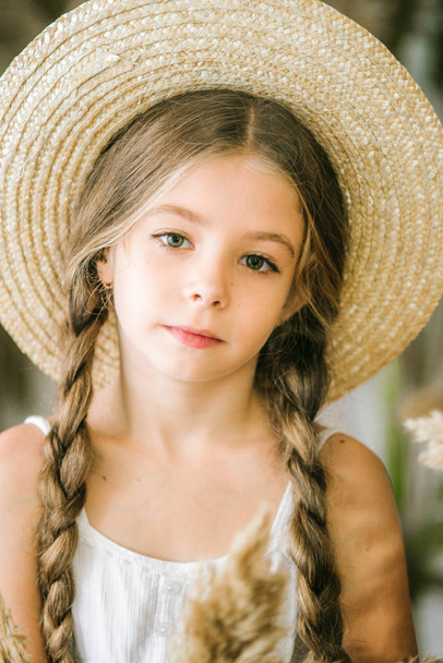 A sweet little girl with long blond hair in a white sarafan and a straw hat in a rattan chair and a reed decor - Photo, image