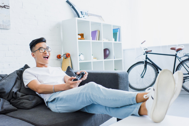 возбужденный красивый азиатский мужчина играет в видеоигры на диване в гостиной
 - Фото, изображение