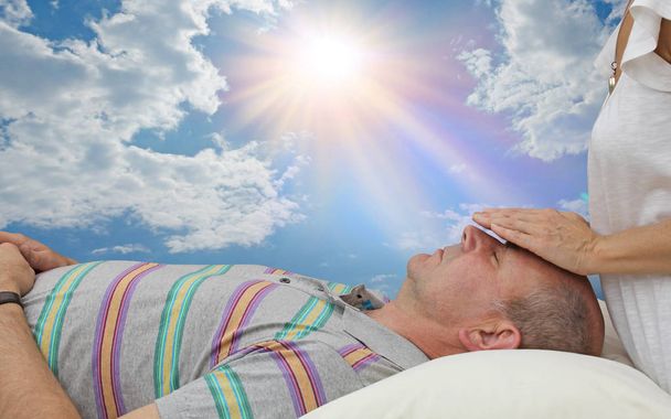 Проведение энергетического лечения вне дверей - женщина с руками, парящими над лбом человека, лежащими на спине, на красивом небе и солнечном фоне
  - Фото, изображение