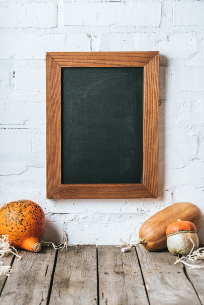 close up view of raw pumpkins on wooden tabletop and blank chalkboard hanging on white brick wall - Photo, Image